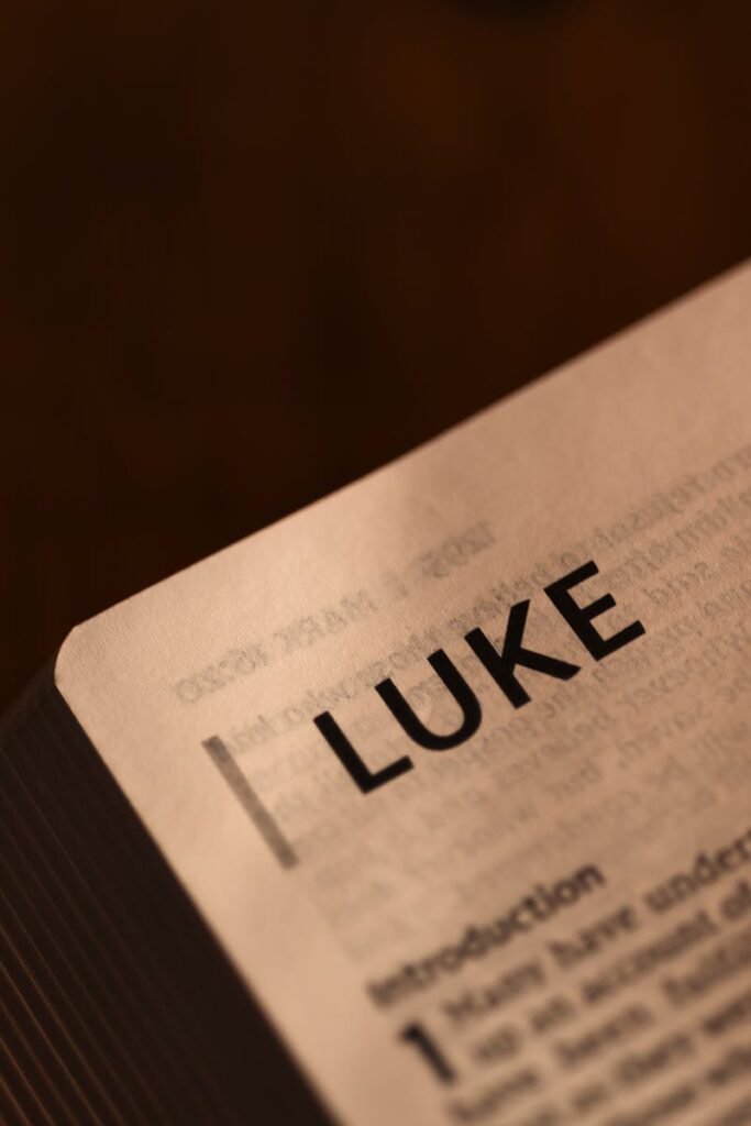 Close-up of an open Bible showing the start of the Book of Luke, softly lit on a dark background.